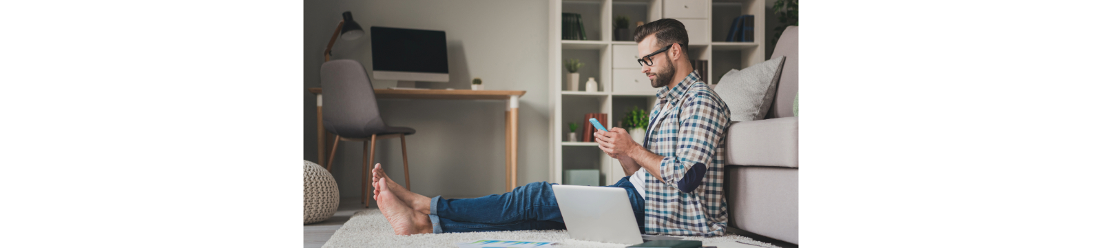 Man in plaid shirt sitting on floor looking at mobile phone with laptop by his side.