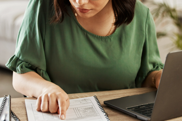 A woman in a green shirt works on her laptop while pointing to a financial worksheet, with a calculator placed beside her.