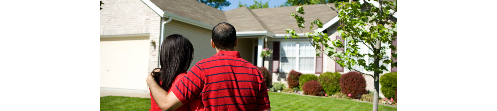 Man with arm around woman looking at the outside of a brick ranch house. 