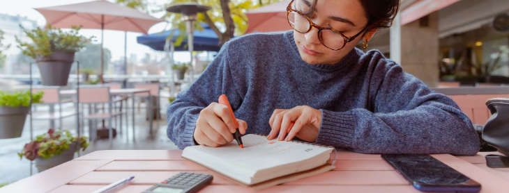 Young woman wearing glasses, sitting at an outdoor table, writing in a notebook with a calculator and smartphone nearby, focused on financial planning.